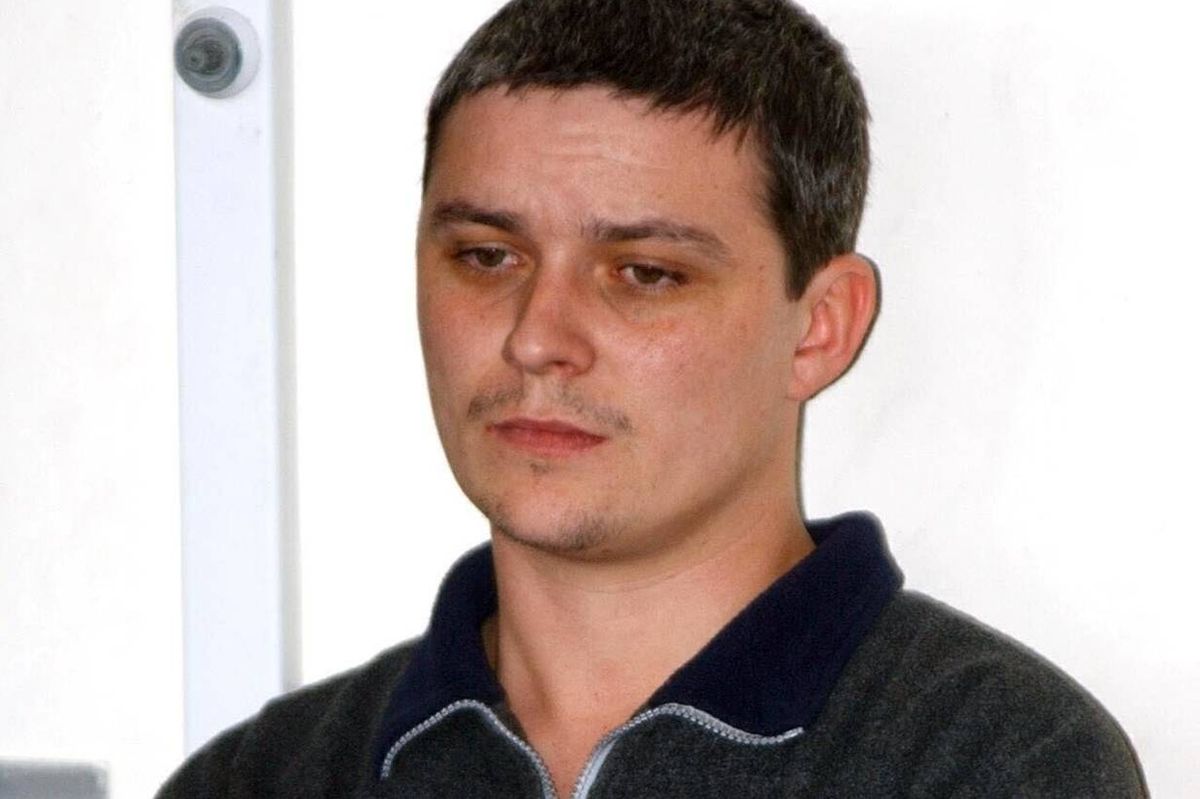 An individual with short, dark hair wearing a black shirt with a blue collar is standing in front of a white wall. The person appears to be in a room, possibly indoors, with focused attention directed towards the camera.