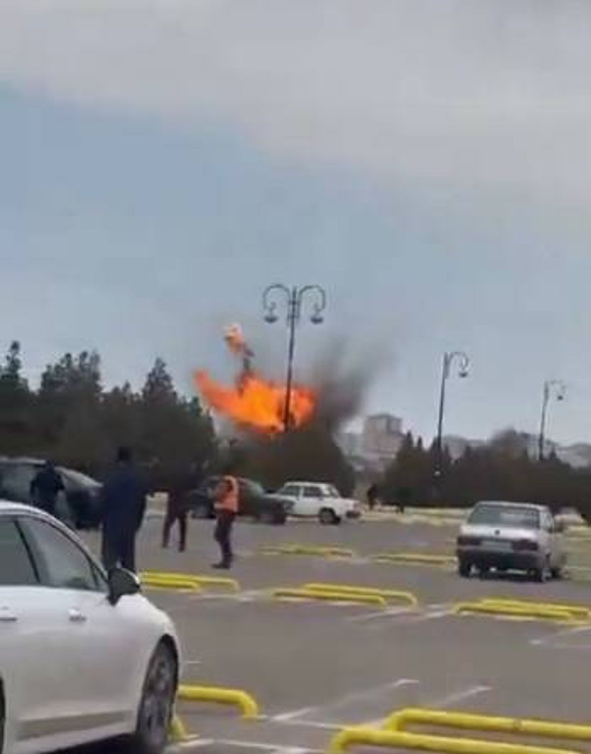 An image depicting an outdoor area with several vehicles parked. A fiery explosion is occurring in the center, amidst a backdrop of trees and streetlights. Several individuals are visible, possibly responding to the incident. The scene is set during the daytime, with a clear sky overhead.