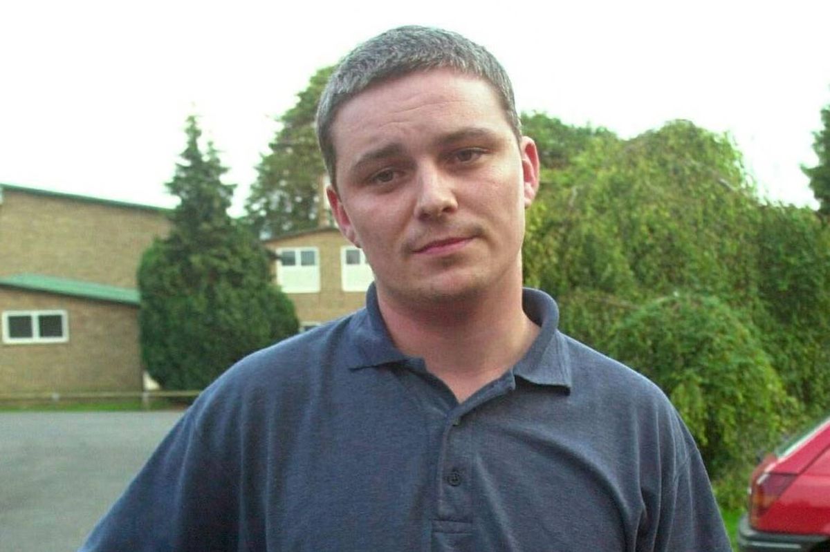 An individual is standing in an outdoor setting, wearing a blue polo shirt. The backdrop features a building with windows and a red car parked nearby, surrounded by greenery.
