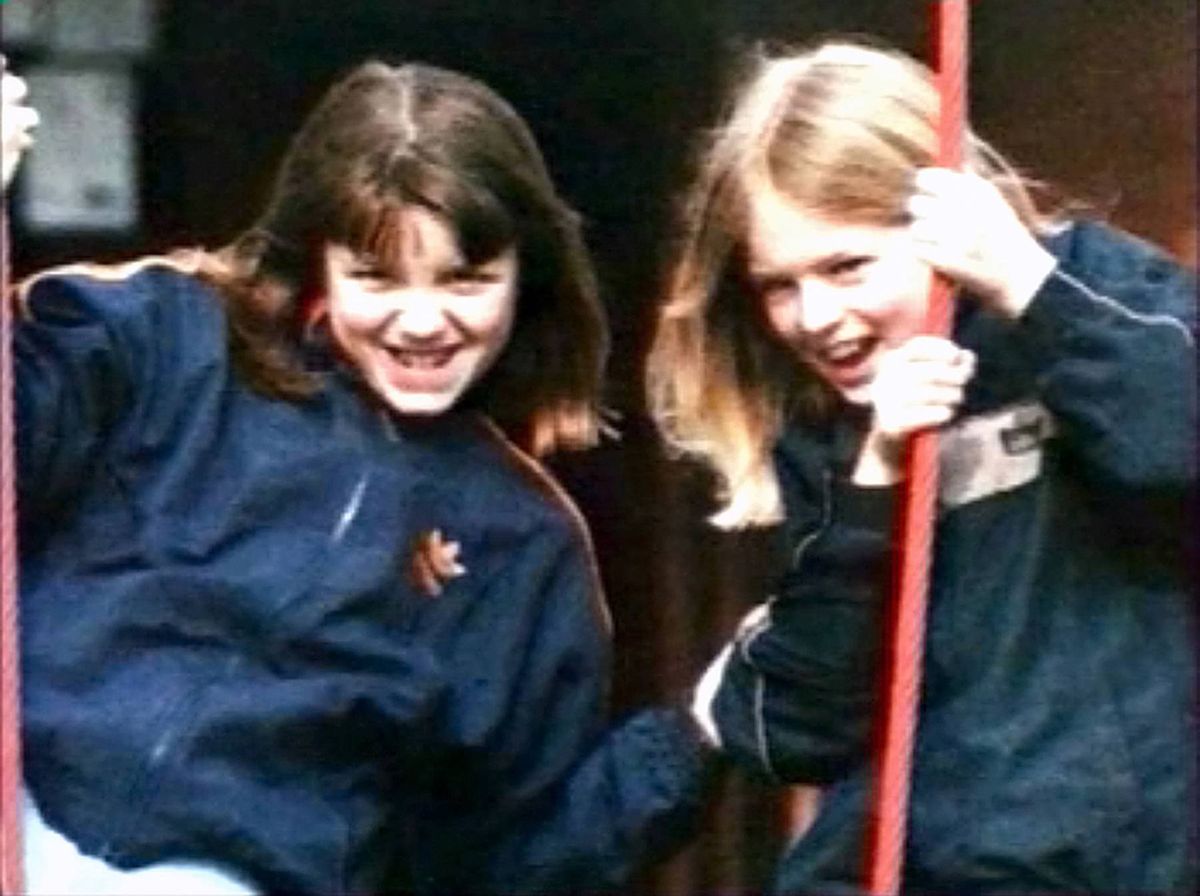 Two individuals dressed in matching blue jackets are joyfully gripping a red pole, seemingly engaged in an activity that elicits their amusement. The scene is set against a backdrop that includes a wall and a sign.