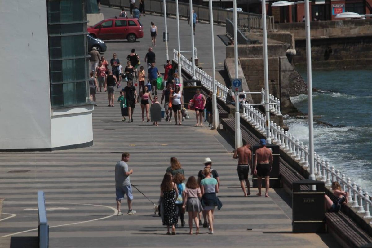 An image depicting a bustling pier with numerous individuals walking along its length, adjacent to a body of water. The pier is equipped with railings and streetlights, and in the background, there is a modern architectural structure.