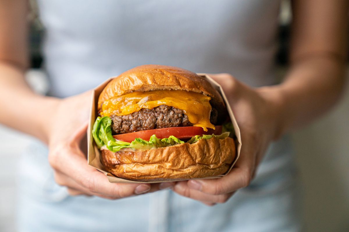 Close up of woman's arms holding a burger to the camera. Beef patty with melted cheese on top, and a large sliced tomato and lettuce at the bottom.