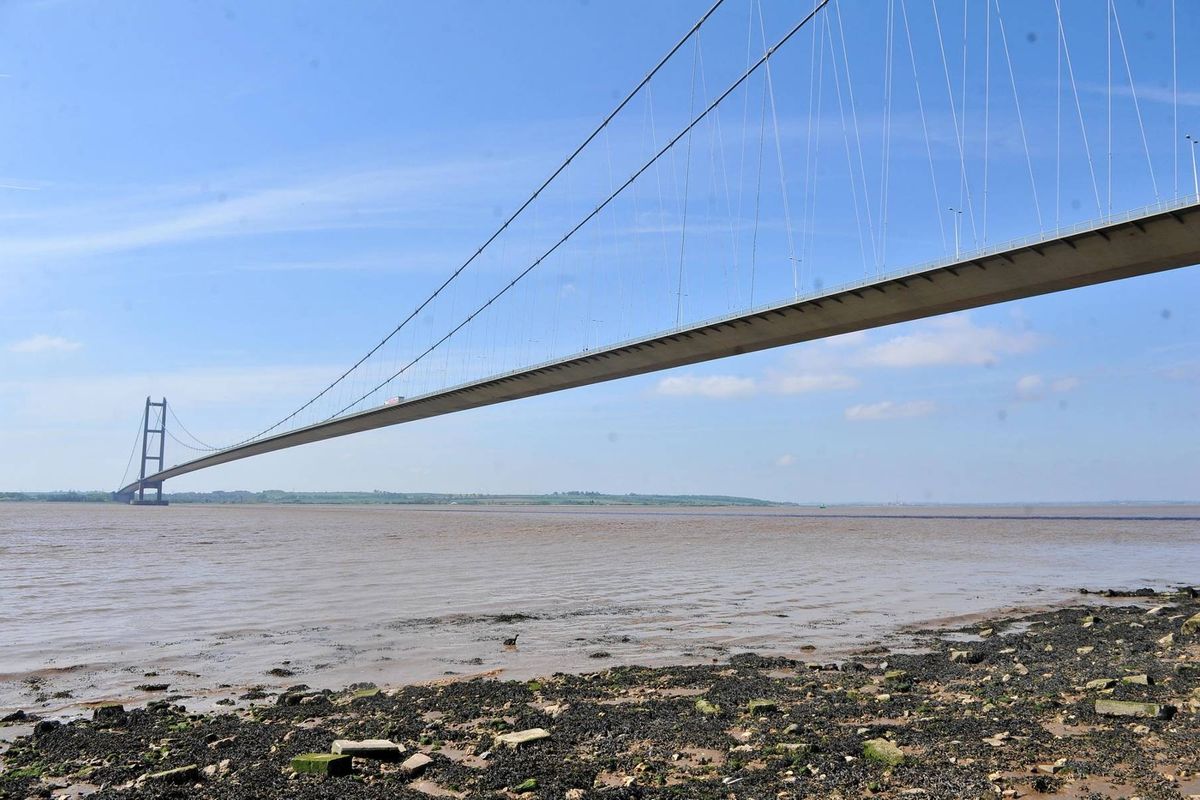 An expansive aerial view of a suspension bridge spanning a body of water, with the clear blue sky in the background and rocky shorelines visible at the foreground.