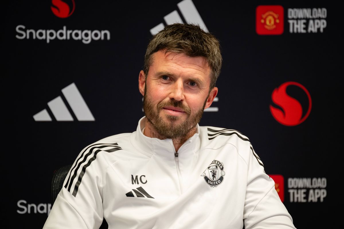 An individual in a white athletic uniform, likely a coach or team member, is seated and addressing the media in front of a backdrop featuring various brand logos.