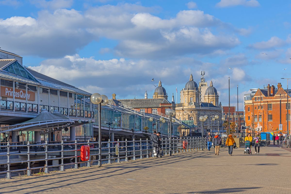 Hull, UK. February 28, 2025. A street scene in a city with a modern glass shopping centre in the foreground. People are on the broad pavement and the towers of a historic building are on the skyline.