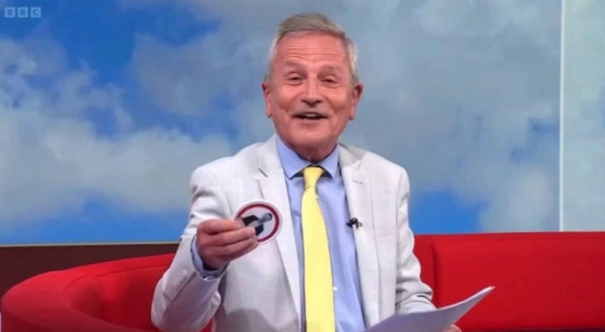 An individual dressed in a white suit with a yellow tie is seated on a red couch, holding a paper and an object in his hand while appearing to speak on a television show.