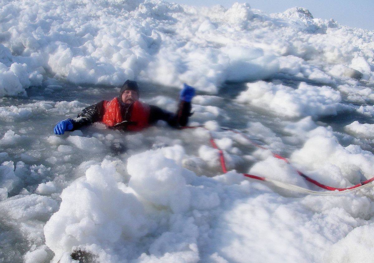 An individual equipped with a life jacket and gloves is lying on their stomach in icy water, attached to a red rope.