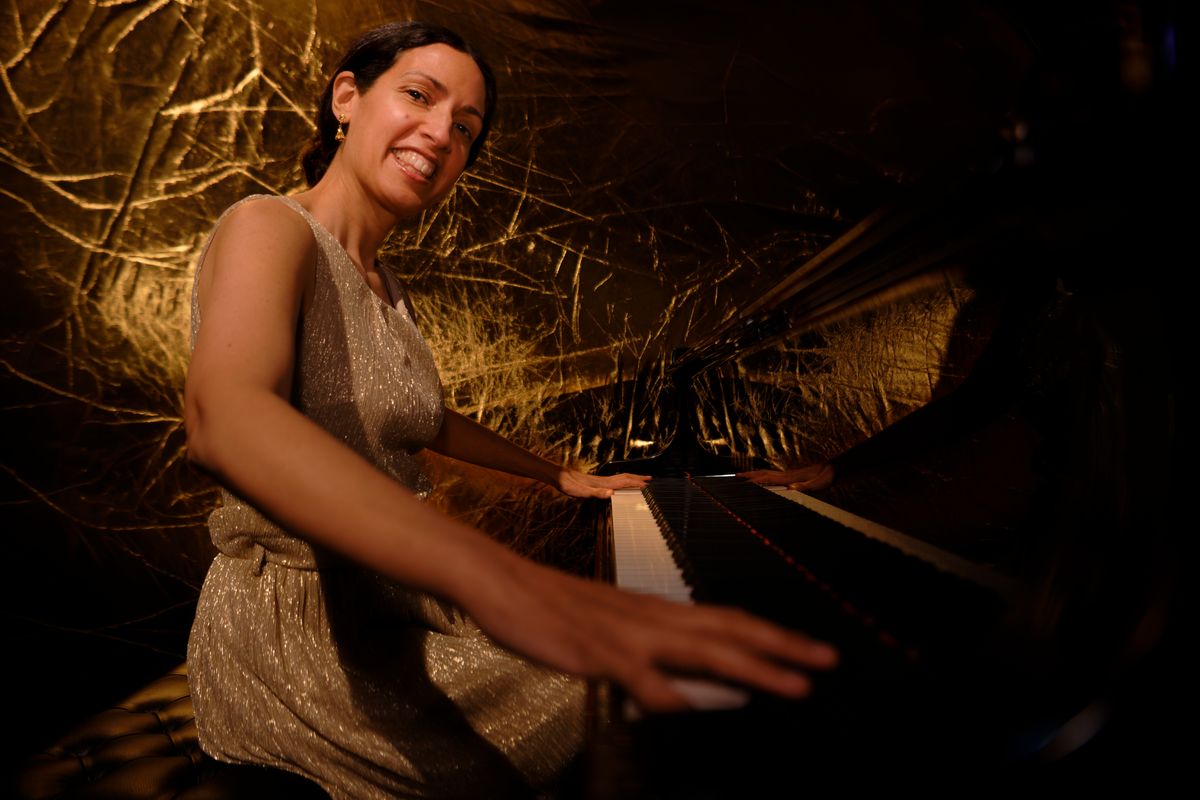Zoe Rahman sits at a piano with her hands on the keys while she smiles towards the camera.