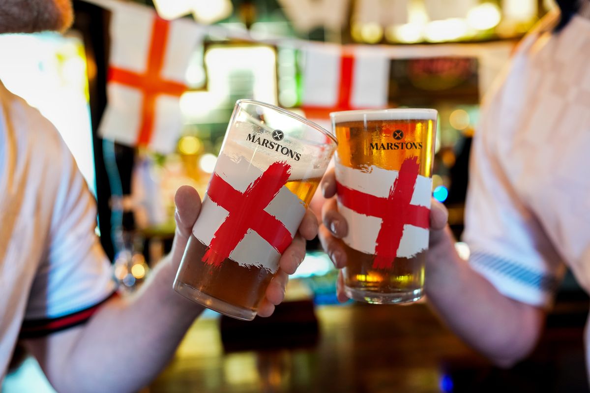 Fans cheers inside a pub using the Marston's promotional England flag pint glasses, one half filled with beer, the other full