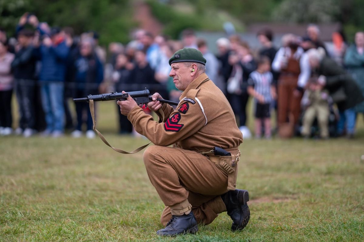 A man in military uniform holds a military weapon at Fort Paull Battery Heritage Site, Holderness, crouched on the ground while crowds watch in the background