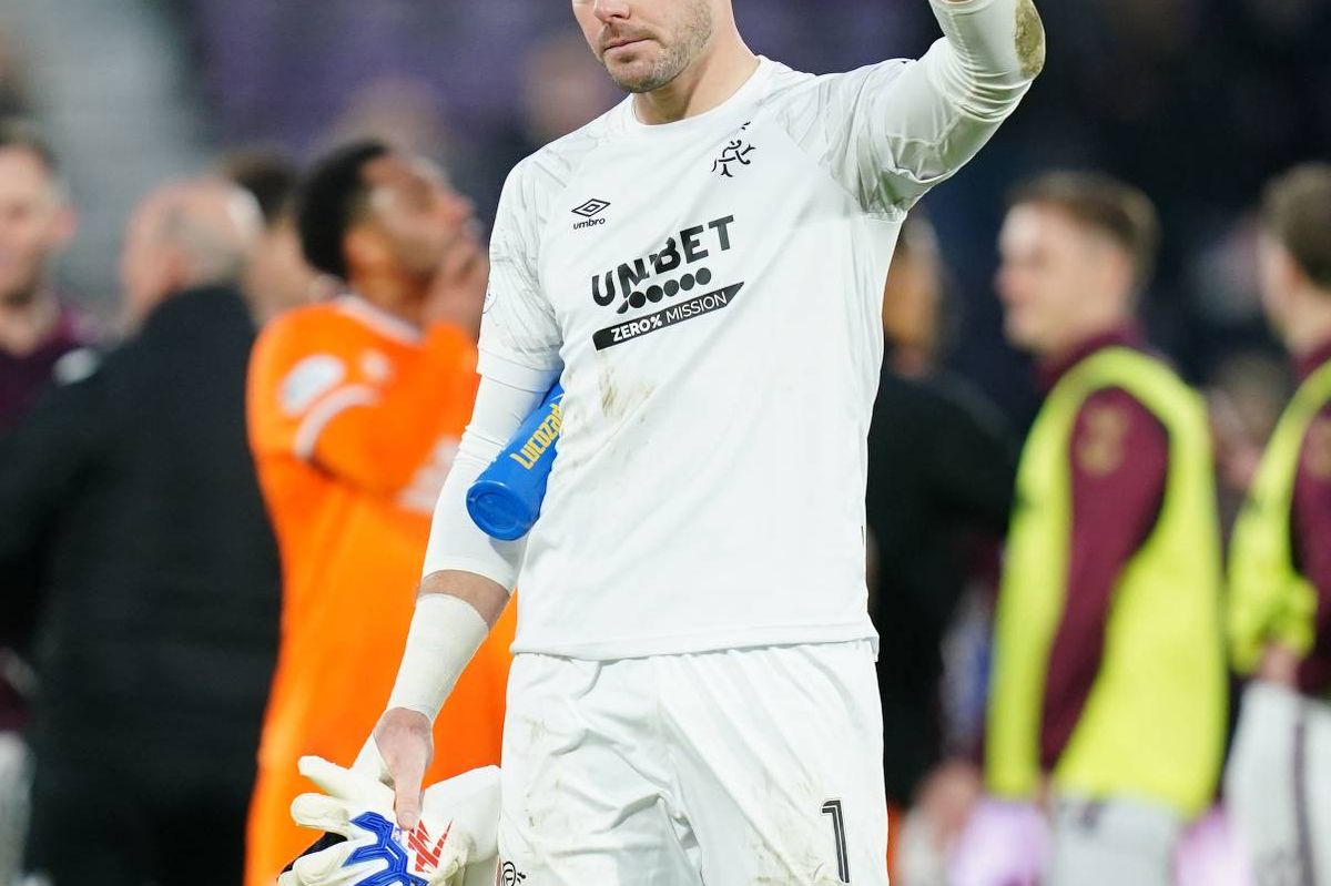 A male athlete in a white jersey waves to the crowd as he walks off the field, with spectators in the background.