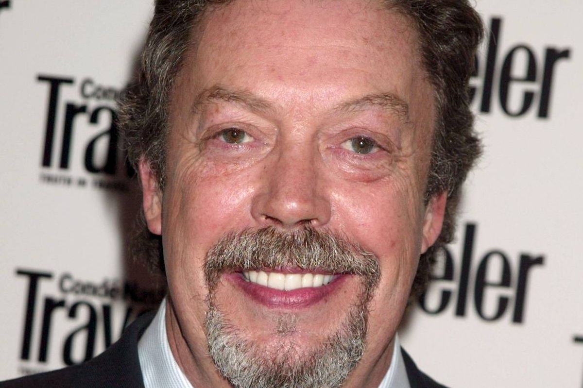 An individual with a beard and mustache, wearing formal attire, is smiling warmly for a photograph against a backdrop featuring the word "Tra."